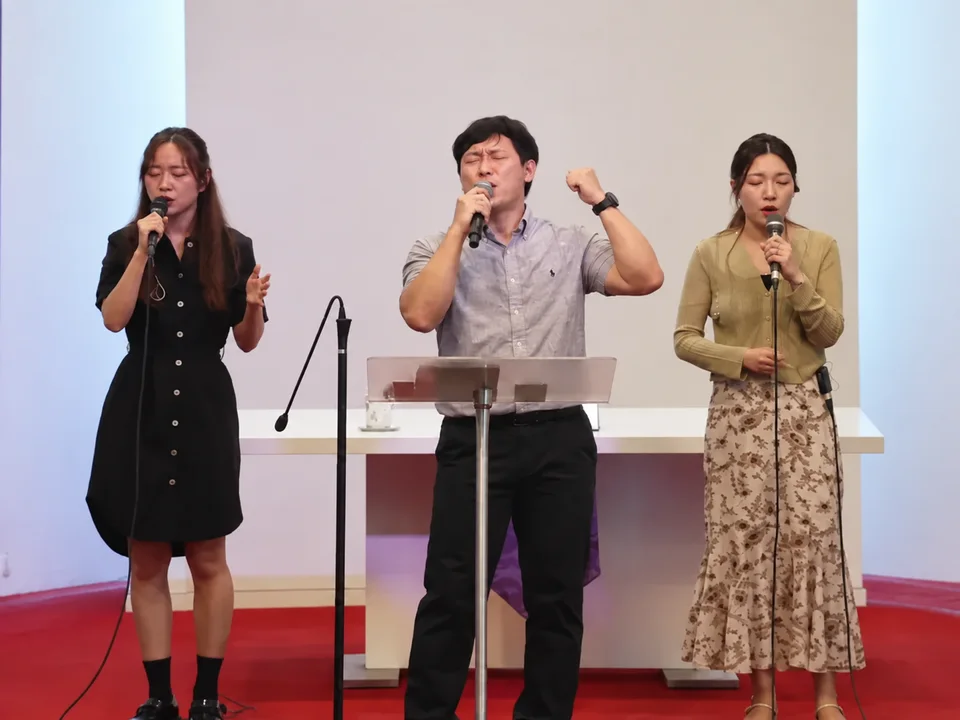 교회 무대에서 마이크를 들고 찬양하는 세 명의 찬양팀